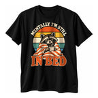 Black t-shirt with a retro sunset, a sleepy raccoon in a striped blanket with a mug and the text Mentally I'm Still In Bed