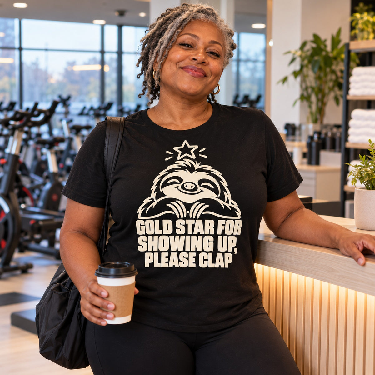 Woman wearing the Line Sloth design black t-shirt at a gym reception holding a takeaway coffee