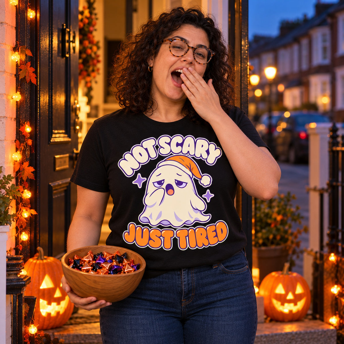 Woman wearing the Sleepy Ghost design black t-shirt on a front doorstep on Halloween evening holding a bowl of sweets