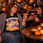 Colleagues wearing the Groovy Witch Cheers design black t-shirt toasting drinks at a pub table with small pumpkins