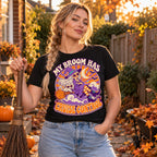 Woman wearing the Retro Witch Badge design black t-shirt holding a straw broom in an autumn garden with pumpkins