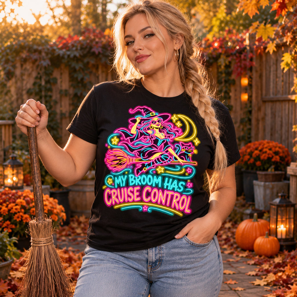 Woman wearing the Neon Witch Cruiser design black t-shirt holding a straw broom in an autumn garden with pumpkins