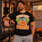 Man wearing the Sticker Strongman Pumpkin design black t-shirt in a home garage gym next to a barbell with pumpkins