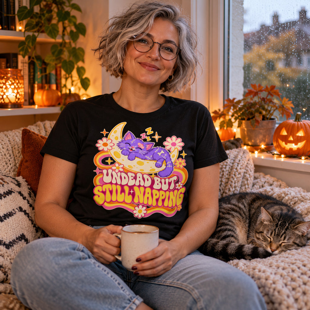 Woman wearing the Groovy Moon Cat design black t-shirt on a cosy window seat with a sleeping cat
