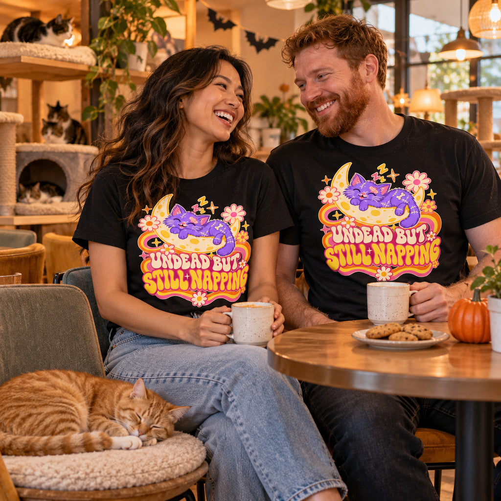 Couple wearing the Groovy Moon Cat design black t-shirts at a cat cafe table with a ginger cat napping