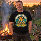 Man wearing the Campfire Badge design black t-shirt at an allotment bonfire at dusk beside a wheelbarrow of branches