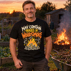 Man wearing the Doodle Campfire design black t-shirt at an allotment bonfire at dusk beside a wheelbarrow of branches