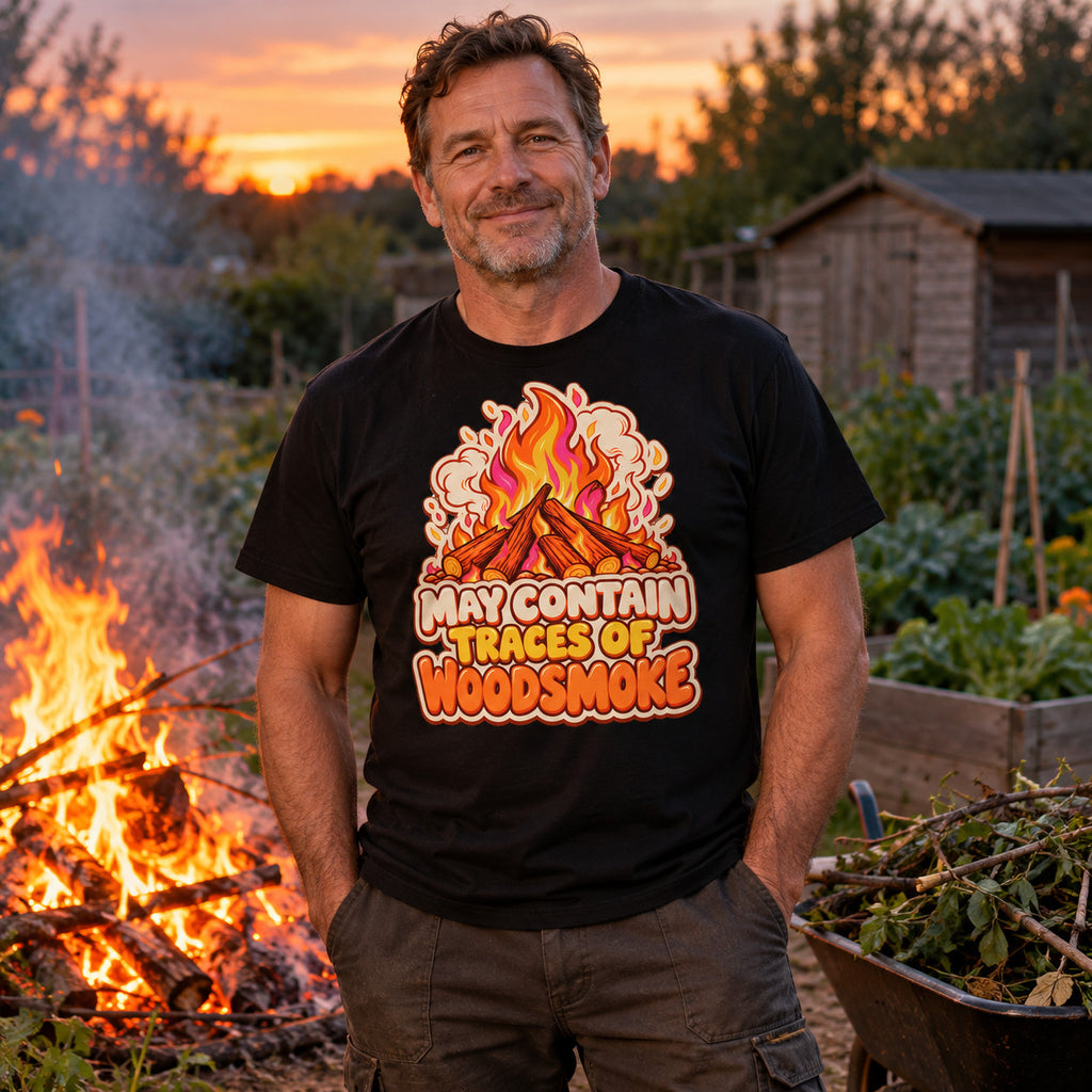 Man wearing the Sticker Campfire design black t-shirt at an allotment bonfire at dusk beside a wheelbarrow of branches