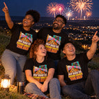 Four friends wearing matching Cartoon Pup design black t-shirts on a hillside at night watching fireworks over the town