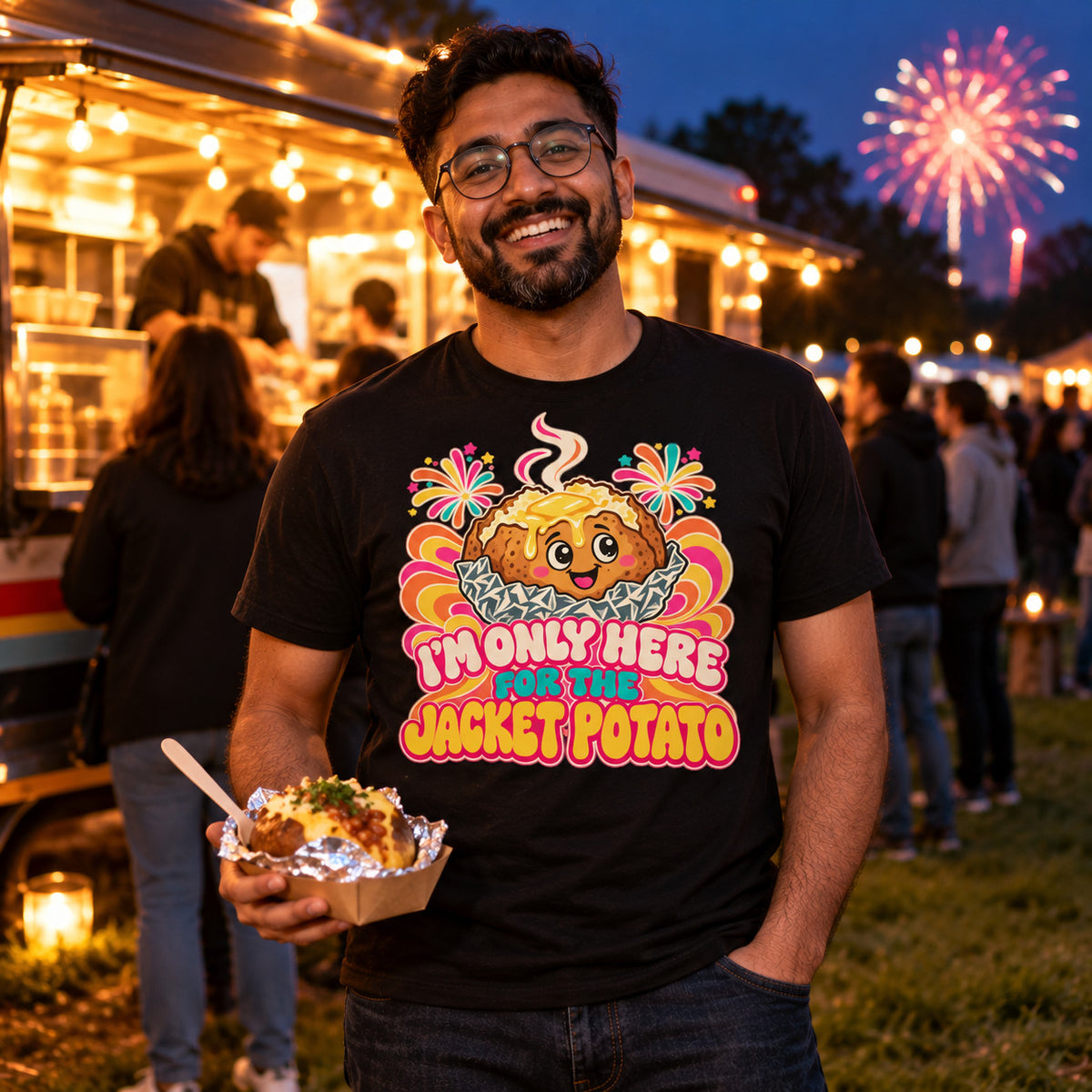 Man wearing the Groovy Spud design black t-shirt at a park street food van at dusk holding a foil wrapped jacket potato with fireworks behind