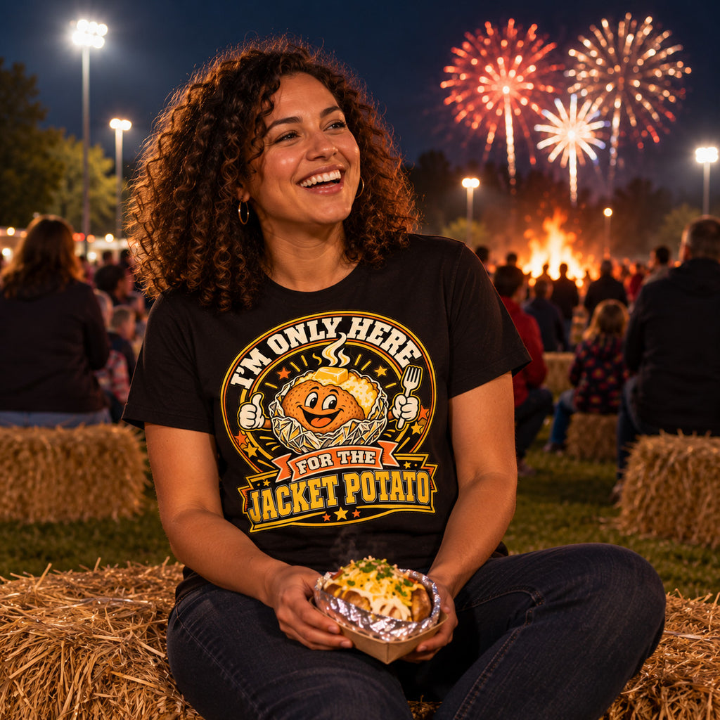 Woman wearing the Doodle Spud design black t-shirt sitting on a hay bale at a school field fireworks night holding a foil wrapped jacket potato