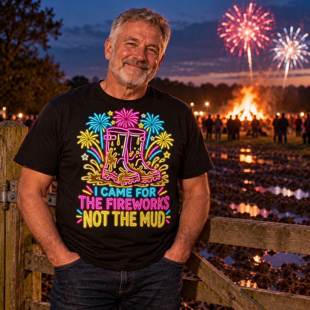 Man wearing the Neon Wellies design black t-shirt at a wooden field gate on a muddy farm at dusk with a bonfire and fireworks behind