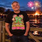 Man wearing the Cartoon Wellies design black t-shirt at a wooden field gate on a muddy farm at dusk with a bonfire and fireworks behind