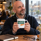 White mug with big stacked deep green capitals and orange italic text reading Working With You Was A Pleasure, Mostly, lifestyle photo 1