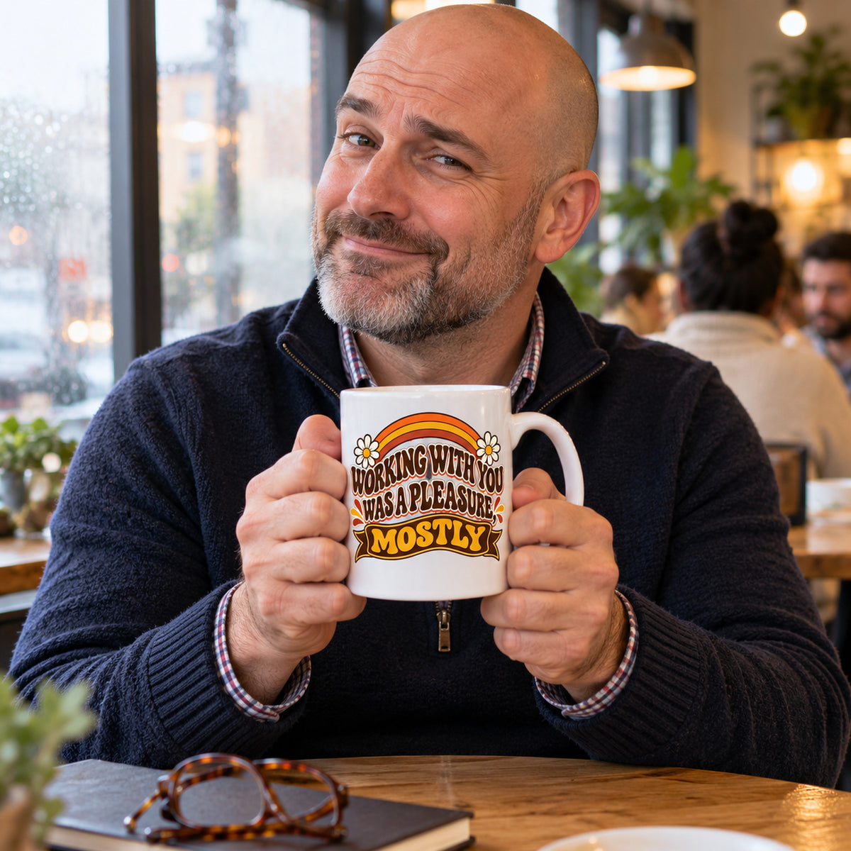 White mug with wavy brown lettering, rainbow arcs, daisies and a banner reading Working With You Was A Pleasure, Mostly, lifestyle photo 1
