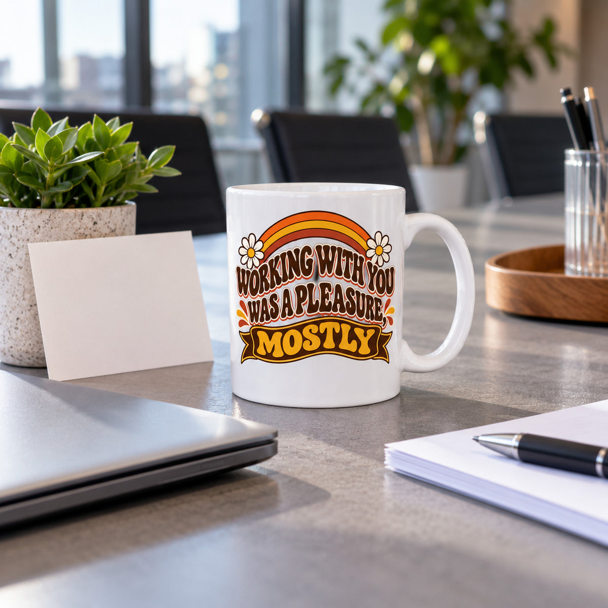 White mug with wavy brown lettering, rainbow arcs, daisies and a banner reading Working With You Was A Pleasure, Mostly, lifestyle photo 2