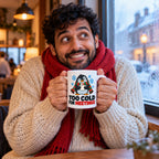 White mug with a grumpy cartoon penguin in a red and green scarf, blue snowflakes and black lettering reading Too Cold For Meetings with a red last word., lifestyle photo 1